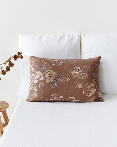 A rectangular decorative pillow with a brown floral pattern, styled on a white bed with white pillows and a small wooden stool with dried leaves on the side.