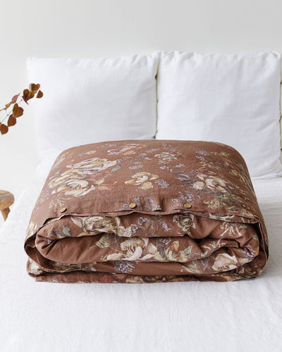A folded brown floral-patterned duvet cover or comforter set with matching pillowcases, styled on a white bed with white pillows and a small decorative plant in the background.