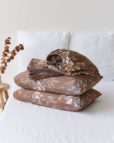 A folded bedding set with a brown floral patterned duvet cover and matching pillowcases, stacked on a white bed with white pillows in the background.