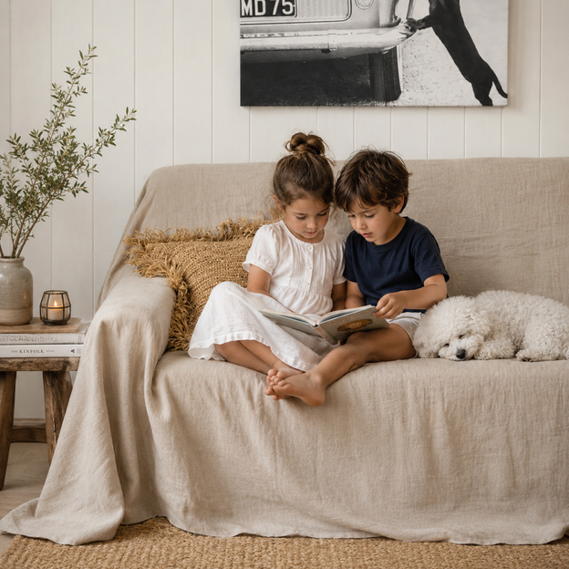 Two children sitting on a beige sofa with a dog, reading a book in a cozy living room.