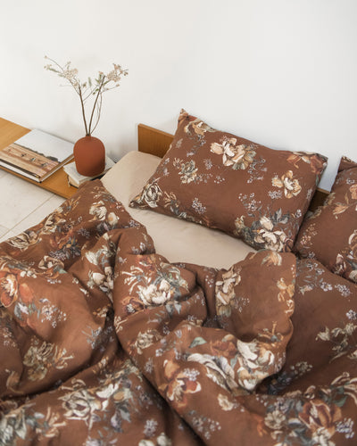 A rectangular decorative pillow with a brown floral pattern, styled on a white bed with white pillows and a small wooden stool with dried leaves on the side.