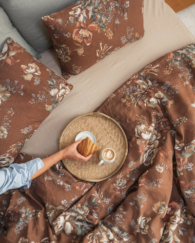 A folded bedding set with a brown floral patterned duvet cover and matching pillowcases, stacked on a white bed with white pillows in the background.