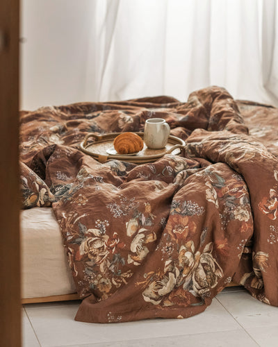 A folded brown floral-patterned duvet cover or comforter set with matching pillowcases, styled on a white bed with white pillows and a small decorative plant in the background.